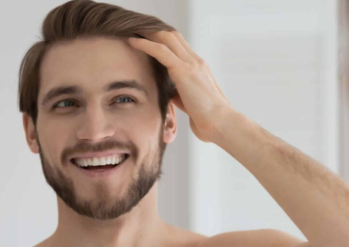Smiling man with a beard touching his hair while looking at his reflection in a mirror.