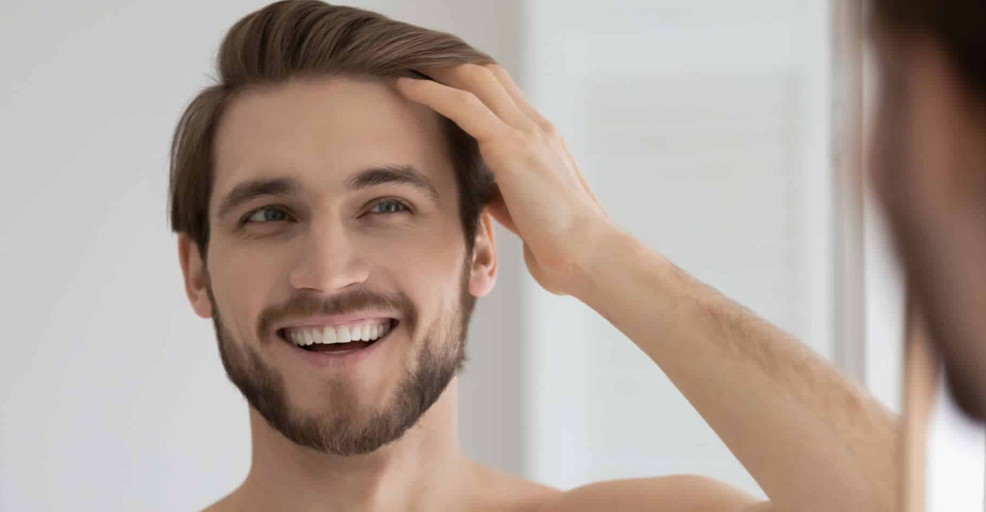Smiling man with a beard touching his hair while looking at his reflection in a mirror.