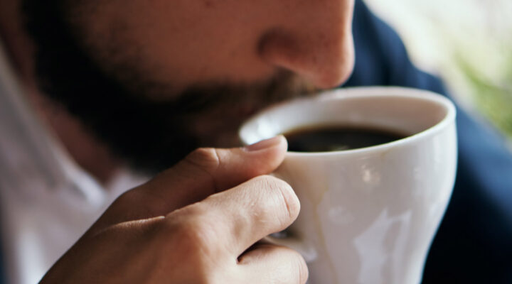 Close-up of a person sipping coffee from a white mug, with a beard and fingers wrapped around the cup.