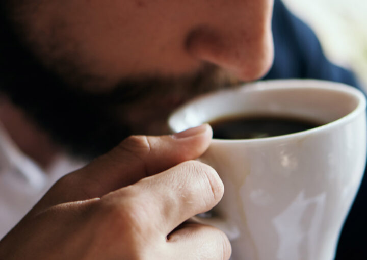 Close-up of a person sipping coffee from a white mug, with a beard and fingers wrapped around the cup.